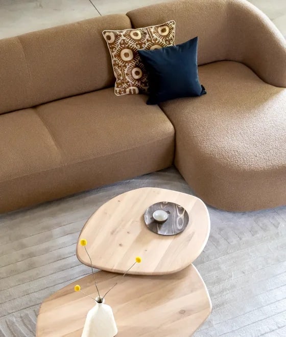 Nahaufnahme eines modularen Sofas in braunem Stoff mit Zierkissen und Couchtischen aus Holz in organischer Form von XOOON